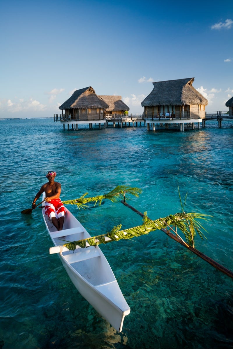 在法属波利尼西亚，一名男子划着独木舟，在清澈的蓝色水面上经过茅草屋顶的房屋